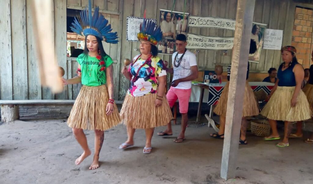 Duas mulheres indígenas Purubora com cocares de penas e saias de palha dançando em assembleia comunitária.