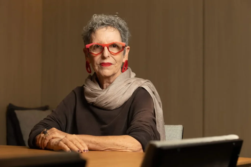 Mulher idosa com cabelo grisalho usando óculos e brincos vermelhos, sentada à mesa.