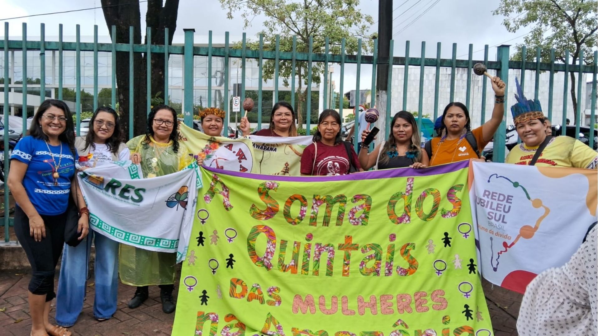 Mulheres em protesto contra violência de gênero em Manaus, incluindo algumas com trajes indígenas, segurando faixas que dizem "A Soma dos Quintais das Mulheres na Amazônia" e "REDE JUBILEU SUL BRASIL".