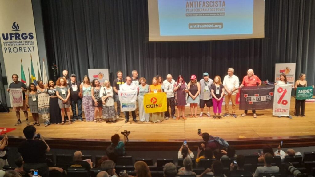 Grupo de ativistas no palco da Conferência Antifascista pela Soberania dos Povos em Porto Alegre, segurando cartazes de organizações como C2RS, CADTM, ATTAC e ANDES durante o encerramento do evento na UFRGS em março de 2026.