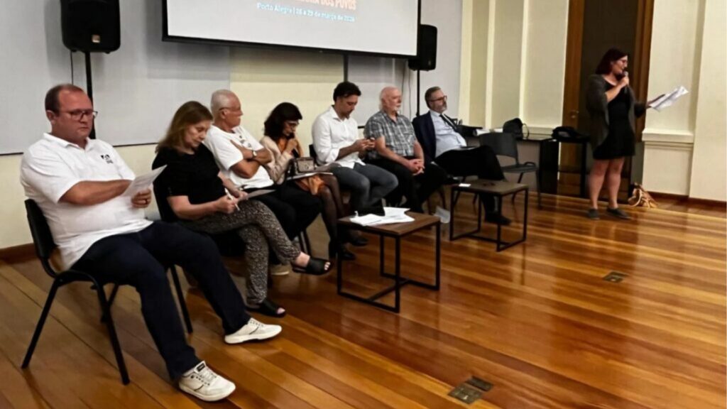 Grupo de pessoas sentadas em palco durante a Assembleia dos Povos em Porto Alegre, de 26 a 28 de março de 2024, com uma mulher falando ao microfone."Este texto descreve o cenário, os participantes principais e o contexto do evento, permitindo que usuários com deficiência visual entendam o que está sendo apresentado na fotografia.