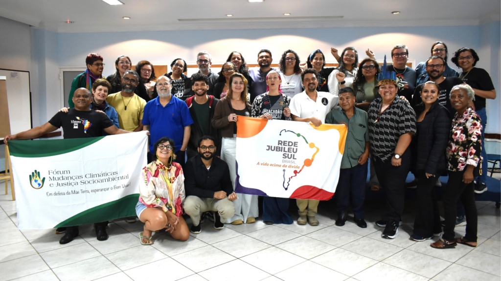 Grupo de ativistas reunidos em evento presencial, segurando faixas do "Fórum Mudanças Climáticas e Justiça Socioambiental" e "Rede Jubileu Sul Brasil - A vida acima da dívida", em ambiente colaborativo para discussão de justiça socioambiental.