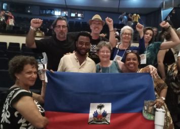 Grupo diverso de pessoas em evento indoor segurando a bandeira haitiana e levantando o punho em gesto de solidaridade.