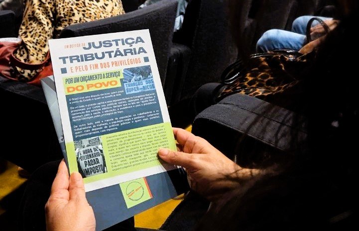 Mãos segurando panfleto de protesto com título "EM DEFESA DA JUSTIÇA TRIBUTÁRIA E PELO FIM DOS PRIVILÉGIOS", exigindo orçamento a serviço do povo e obrigação dos ricos a pagar impostos.