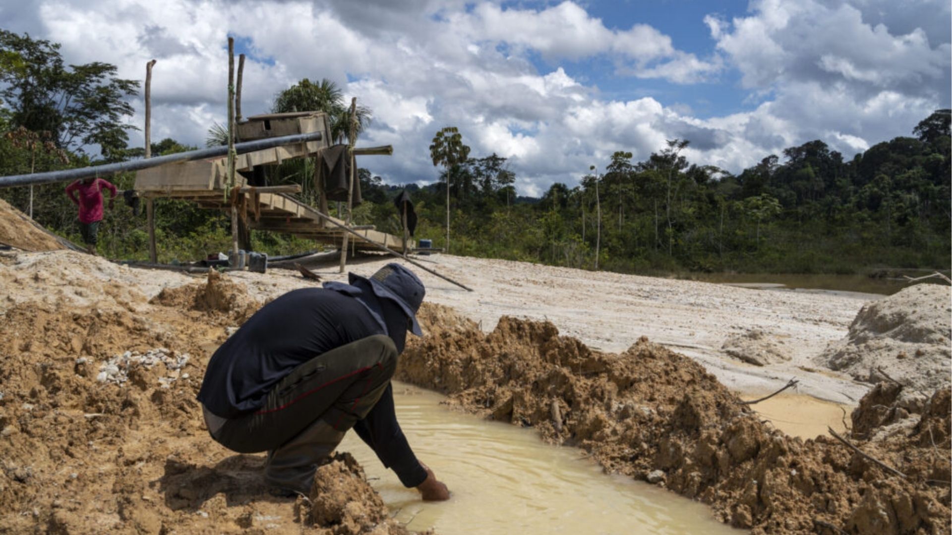Trabalhador em garimpo ilegal manipulando água lamacenta em canal de escavação na Amazônia, com estrutura de mineração ao fundo e floresta degradada ao redor.