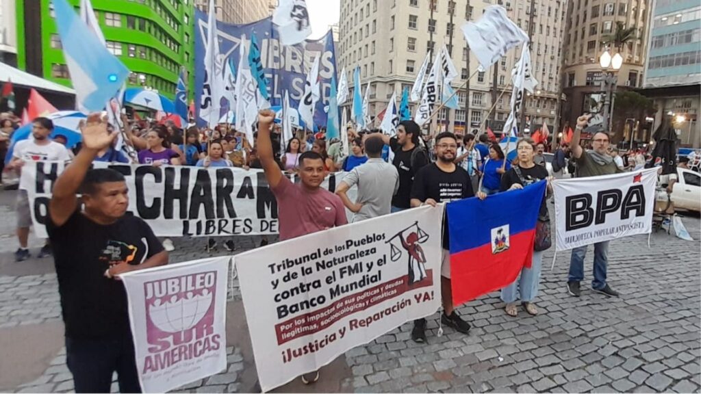 Manifestantes em marcha urbana segurando cartazes exigindo justiça e reparações contra o FMI e Banco Mundial, com destaque para o banner do Jubileo Sur Americas e do Tribunal dos Povos e da Natureza contra as políticas de dívidas ilegítimas.