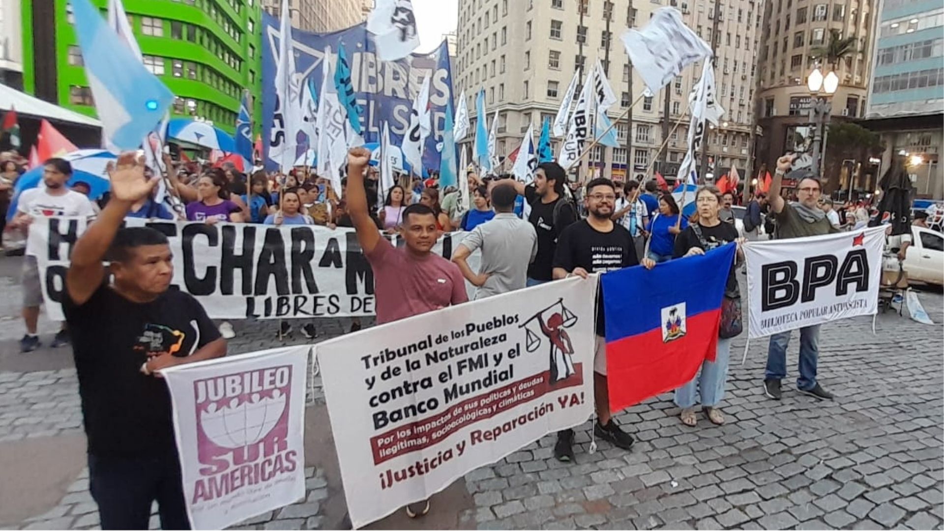 Manifestantes em marcha urbana segurando cartazes exigindo justiça e reparações contra o FMI e Banco Mundial, com destaque para o banner do Jubileo Sur Americas e do Tribunal dos Povos e da Natureza contra as políticas de dívidas ilegítimas.