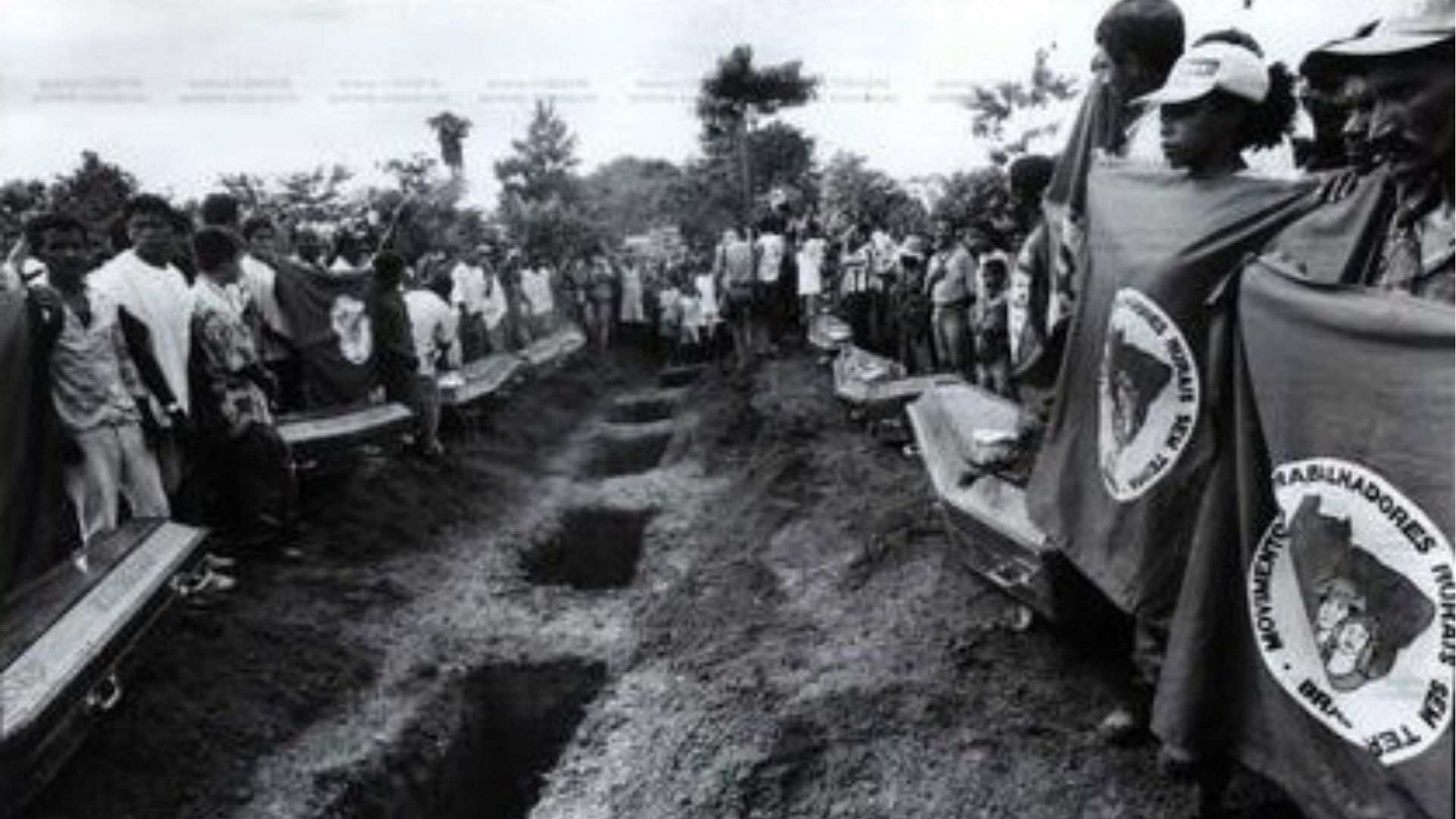 Fotografia histórica em preto e branco mostrando o funeral coletivo das 21 vítimas do massacre de Eldorado dos Carajás em 17 de abril de 1996, com caixões alinhados e multidão ao redor, incluindo militante com bandeira do MST.