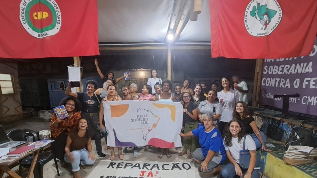 Grupo de mulheres em evento noturno da Rede Jubileu Sul Brasil, segurando faixa central 'A vida acima da dívida', com bandeiras da CMP e MST e faixa no chão com lema 'REPARAÇÃO JÁ!