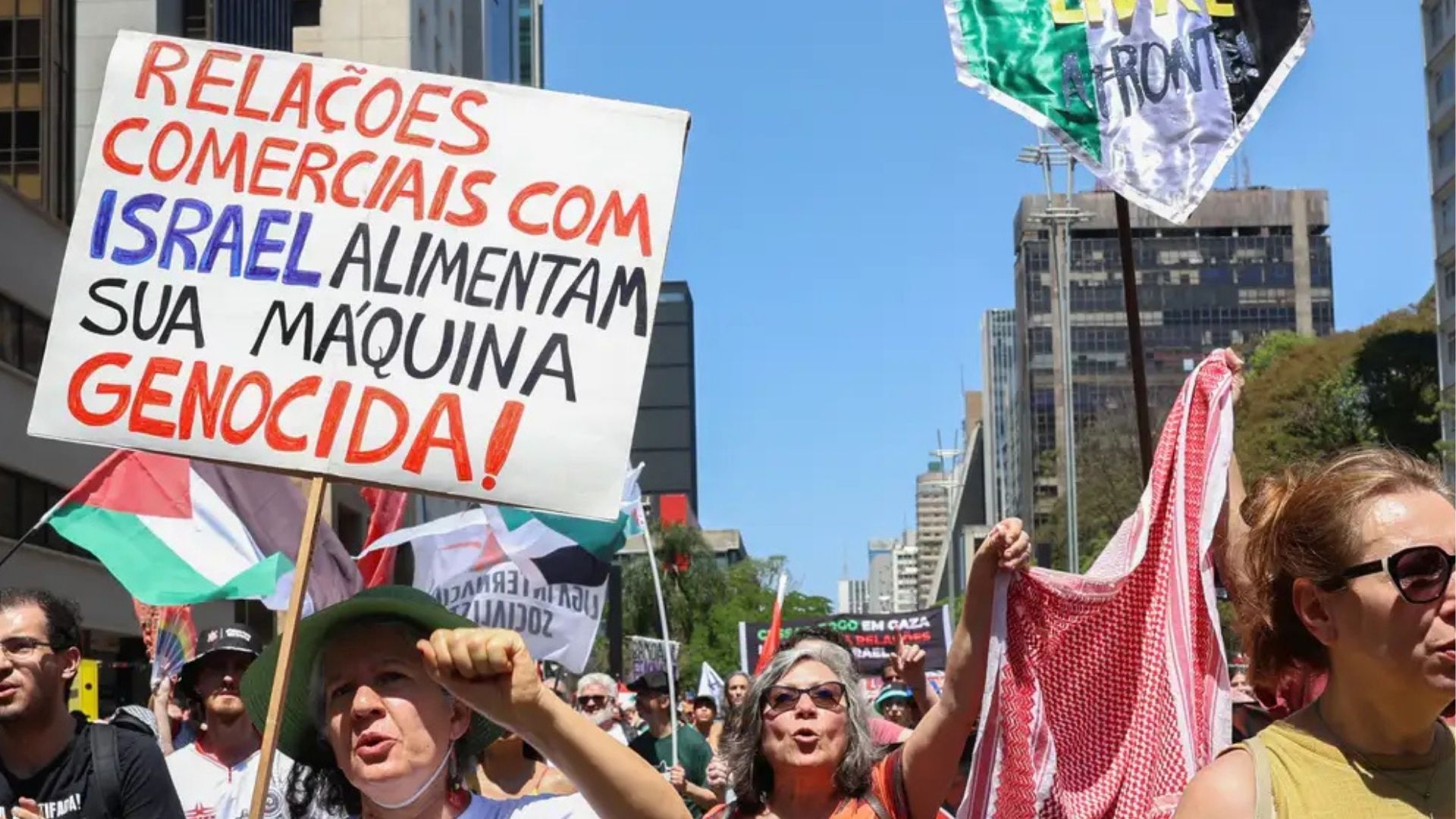 Manifestantes em protesto urbano segurando cartazes e bandeira palestina, com um cartaz destacado que diz "RELAÇÕES COMERCIAIS COM ISRAEL ALIMENTAM SUA MÁQUINA GENOCIDA!".