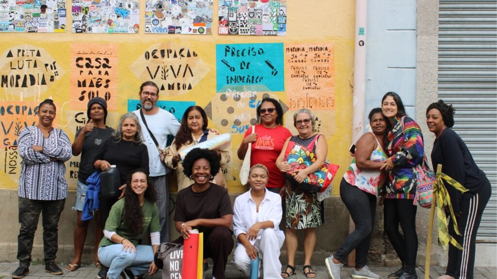 Grupo de pessoas posando em frente a um mural colorido com mensagens de luta por moradia digna. O mural contém frases em português como 'A preta luta por moradia', 'Casa para mãe solo' e 'Preciso de endereço', refletindo o movimento por direitos habitacionais, especialmente da população negra e mulheres