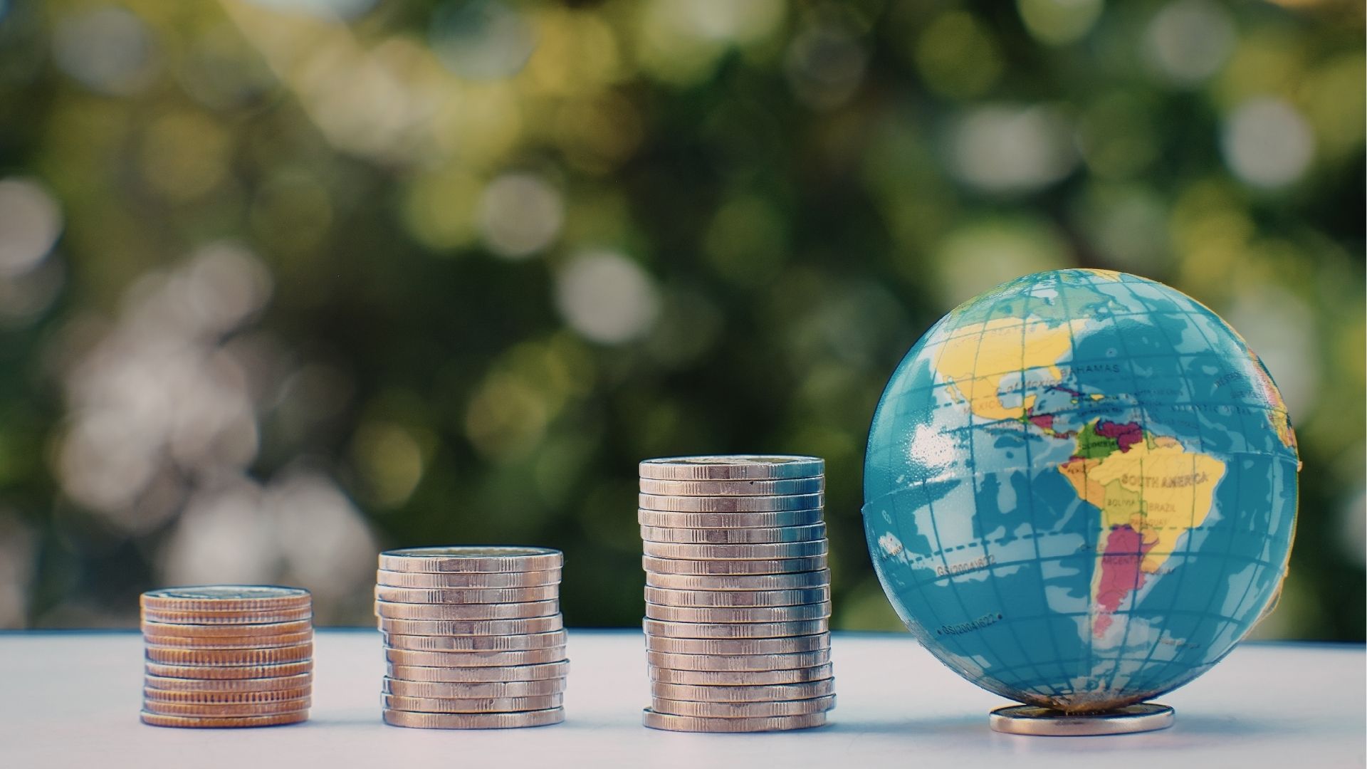 Três pilhas de moedas de diferentes alturas ao lado de um globo terrestre, simbolizando economia global e finanças internacionais.