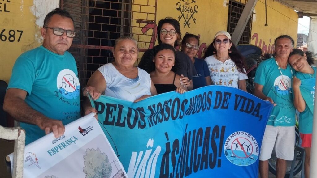Comunidade reunida em roda de conversa, segurando faixa azul com texto "PELOS NOSSOS MODOS DE VIDA, NÃO ÀS EÓLICAS!" e logótipo de turbina eólica riscada sobre cena marinha, em protesto contra parques eólicos marinhos.