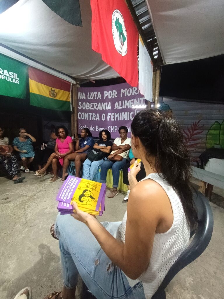 Mulheres em roda de conversa no Rio de Janeiro, participando de oficina temática sobre enfretamento à violência contra a mulher e combate ao feminicídio, com panfletos e bandeiras de movimentos sociais.