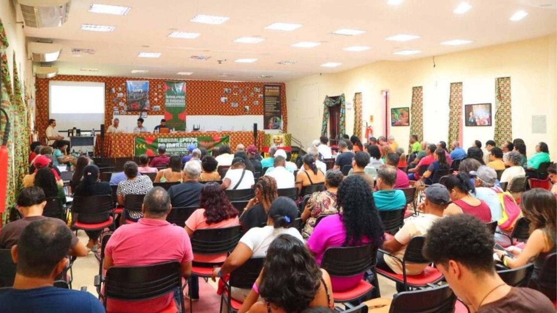 Audiência popular com pessoas sentadas em sala, ouvindo painel de discussão no palco. Banner com texto "PELOS NOSSOS MODOS DE VIDA, NÃO ÀS EÓLICAS!" e projetor exibindo imagem. Ambiente comunitário formal e engajado.