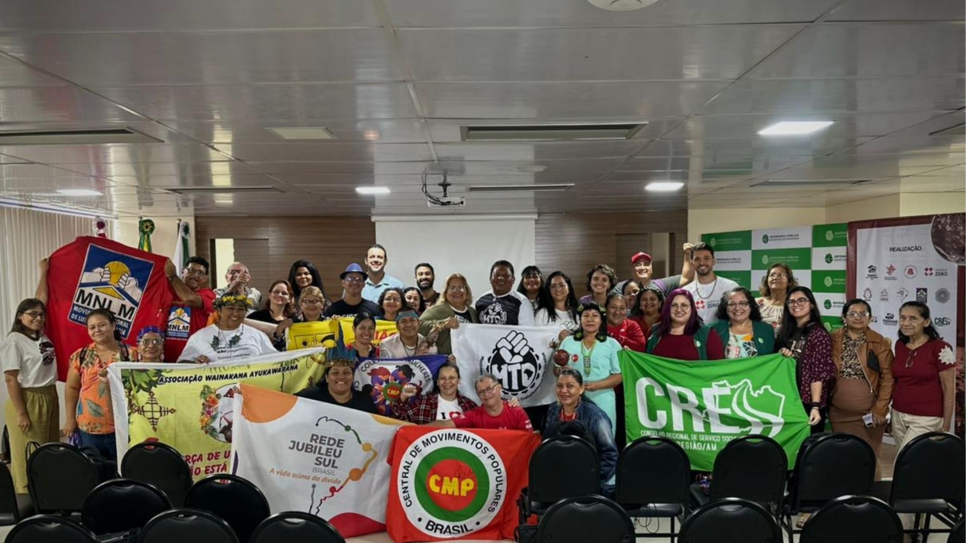 Grupo de comunidades em debate sobre moradia e clima promovido pela Defensoria do Amazonas, segurando bandeiras de organizações sociais e ambientais durante encontro.