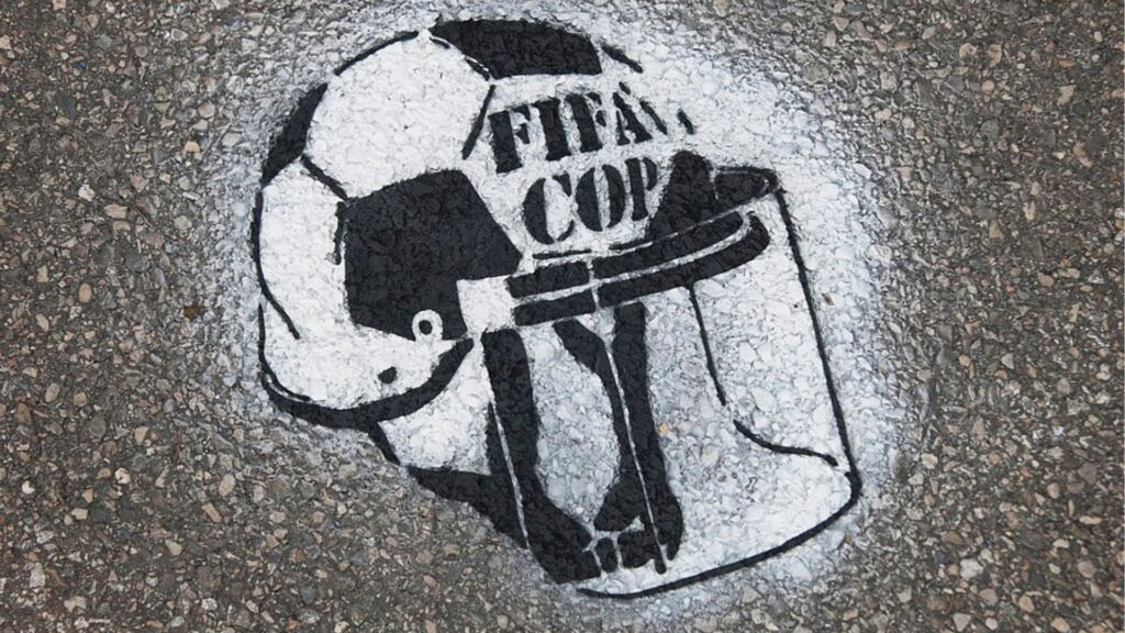 Arte de rua em spray no chão, mostrando um capacete de policial sobre uma bola de futebol com o texto "FIFA COP", criticando a repressão policial durante a Copa do Mundo FIFA.