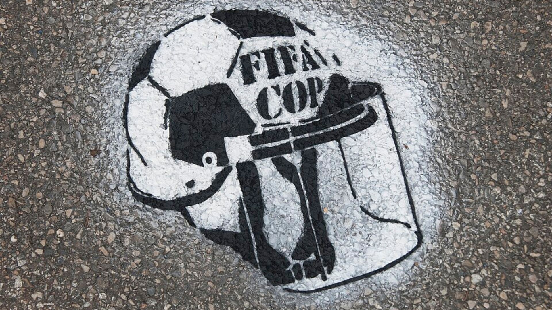 Arte de rua em spray no chão, mostrando um capacete de policial sobre uma bola de futebol com o texto "FIFA COP", criticando a repressão policial durante a Copa do Mundo FIFA.
