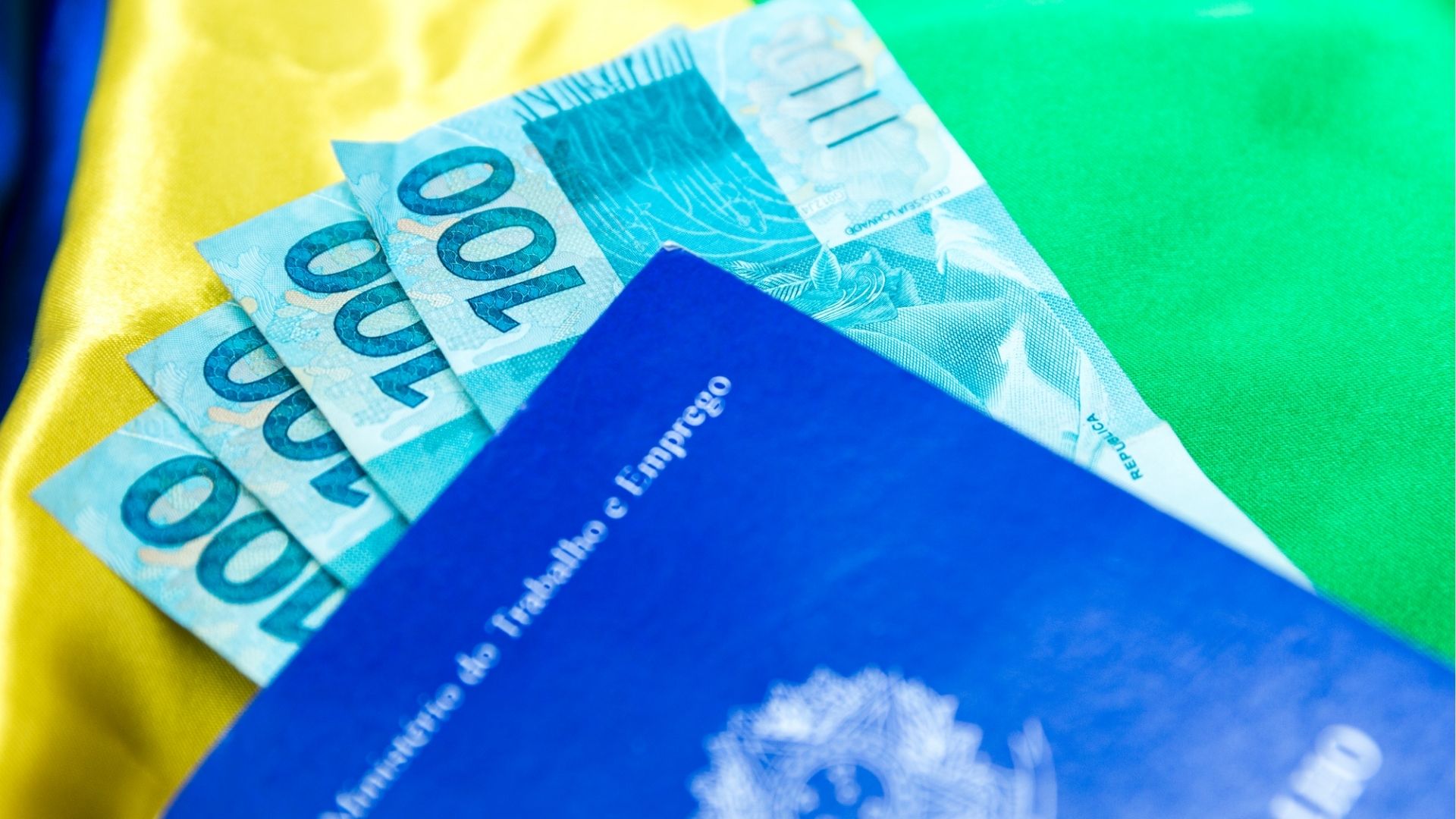 Notas de 100 reais e documento do Ministério do Trabalho e Emprego sobre fundo da bandeira do Brasil.