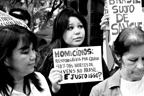 Três mulheres em protesto, com destaque para a central que segura cartaz em português: "Homicídios: responsáveis por quase 40% das mortes de jovens no Brasil. E justo isso?"