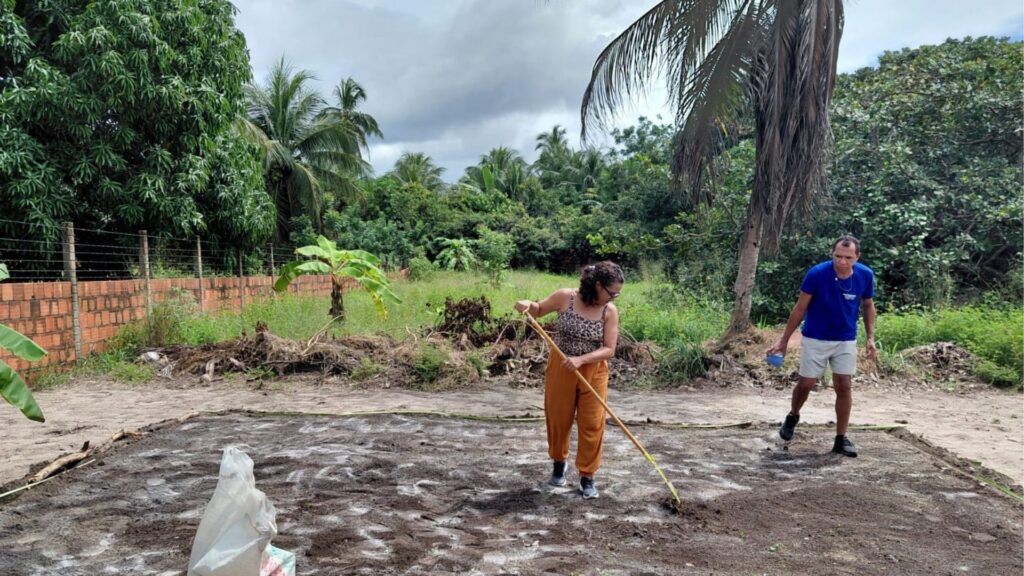 Duas pessoas trabalhando em horta comunitária em Paraíbapa, mulher usando bastão para cultivar solo e homem segurando recipiente, cercada por vegetação densa e muros de tijolos.