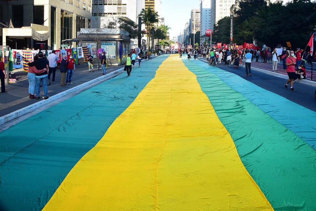 Manifestação na Avenida Paulista com grande faixa colorida estendida no asfalto, pessoas carregando bandeiras e placas em evento coletivo urbano.