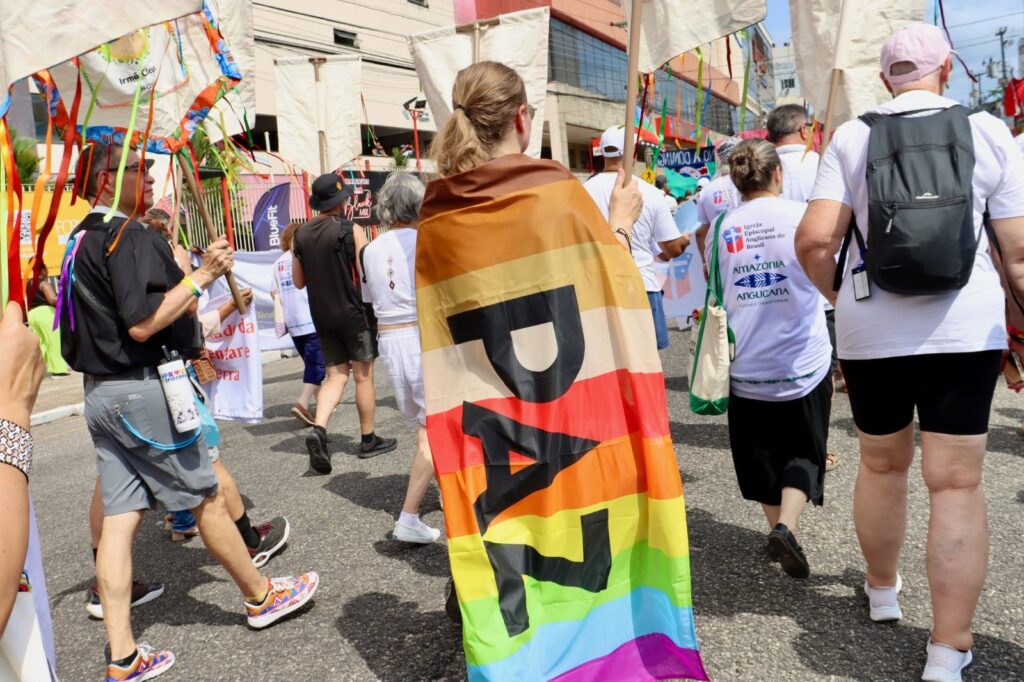 escreva o texto alternativo curto da imagem 3:55 PM Manifestação LGBTQI+ com pessoas marchando e carregando bandeiras. Destaque para pessoa com faixa colorida que diz "JUSTIÇA" em português, em protesto ou celebração.