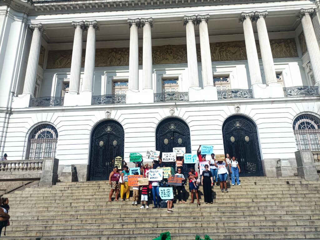 Mulheres e crianças protestam nas escadarias de um edifício governamental neoclássico com colunas brancas, segurando cartazes coloridos exigindo moradia digna e direitos sociais.