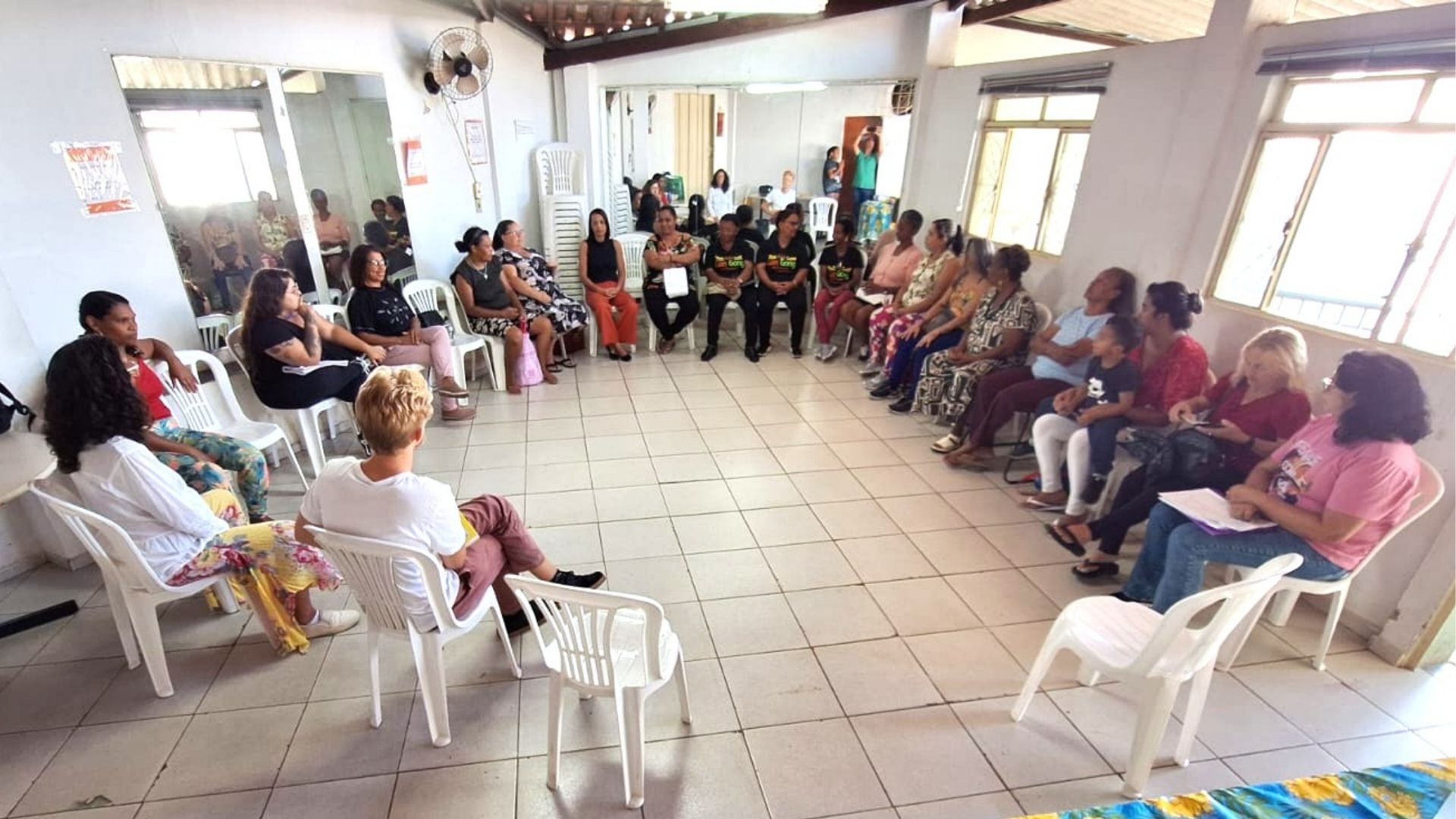Grupo de pessoas sentadas em cadeiras de plástico brancas formando uma roda de conversa em um salão amplo com piso de azulejos.