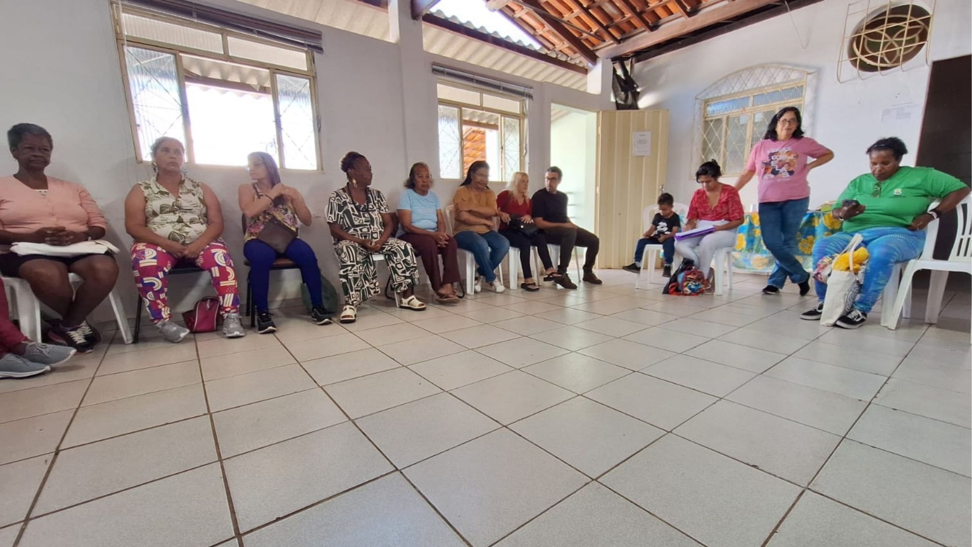 Grupo de pessoas em oficina de autocuidado em Belo Horizonte, sentadas em círculo em sala com piso azulejado e grandes janelas.