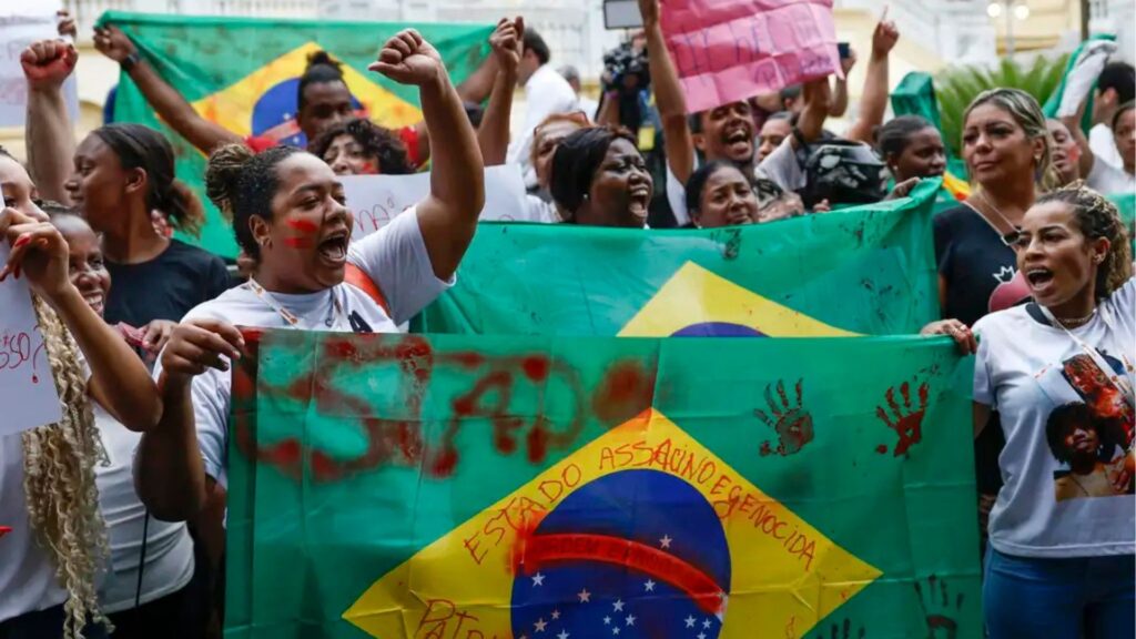 Mulheres negras protestam em rua urbana, segurando bandeira do Brasil modificada com as palavras "ESTADO ASSASSINO E GENOCIDA" e marcas de mãos vermelhas. Manifestantes gritam com punhos erguidos em ato contra violência estatal.