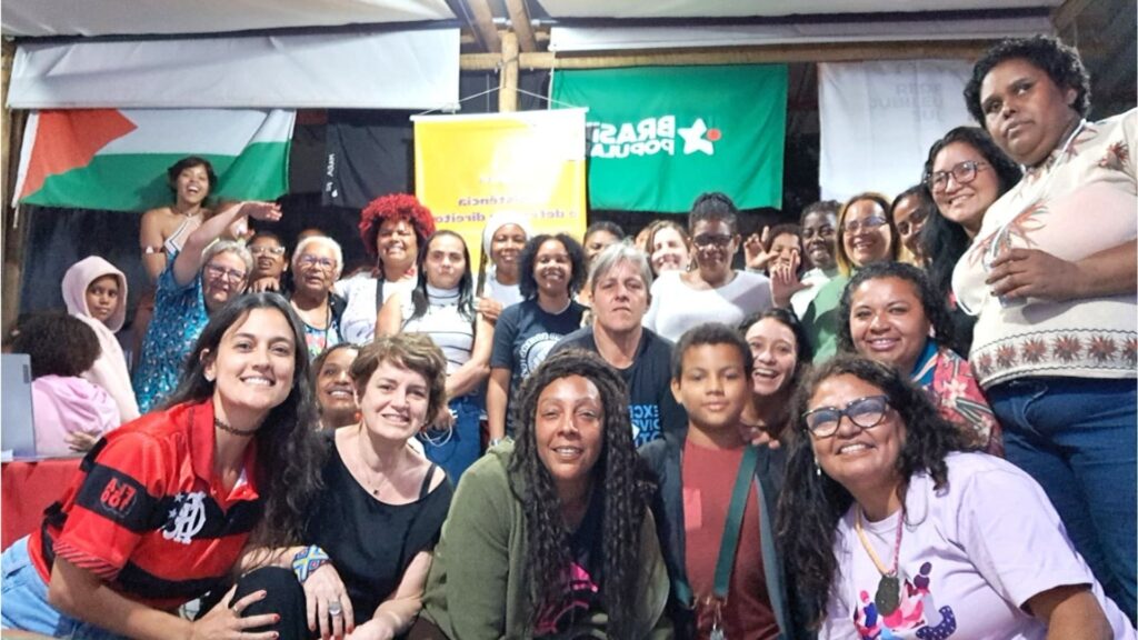 Grupo diverso de pessoas sorrindo em evento ao ar livre, com bandeiras do Brasil Popular e Palestina ao fundo. Mulheres negras estão em destaque na frente, algumas sentadas e outras em pé, num ambiente com tendas e faixas políticas.