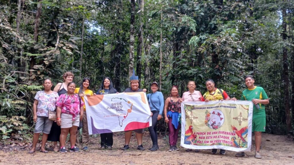 Grupo de pessoas em floresta segurando bandeiras da Rede Jubileu Sul e Associação Wainakana Ayukawarana, com a mensagem "A força de uma guerreira não está no ataque, mas sim na resistência".