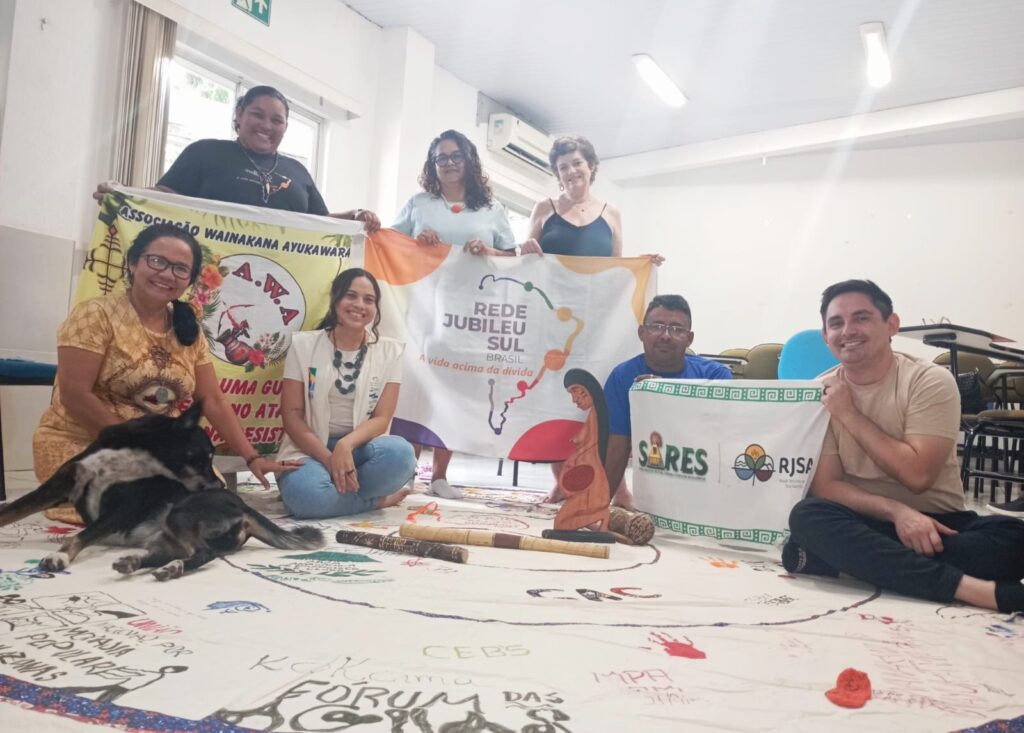 Grupo de pessoas segurando bandeiras da Associação Wainakana Ayukawara, Rede Jubileu Sul Brasil e SARES/RJSA, sentadas em volta de uma grande tela com desenhos e mensagens sobre direitos populares.