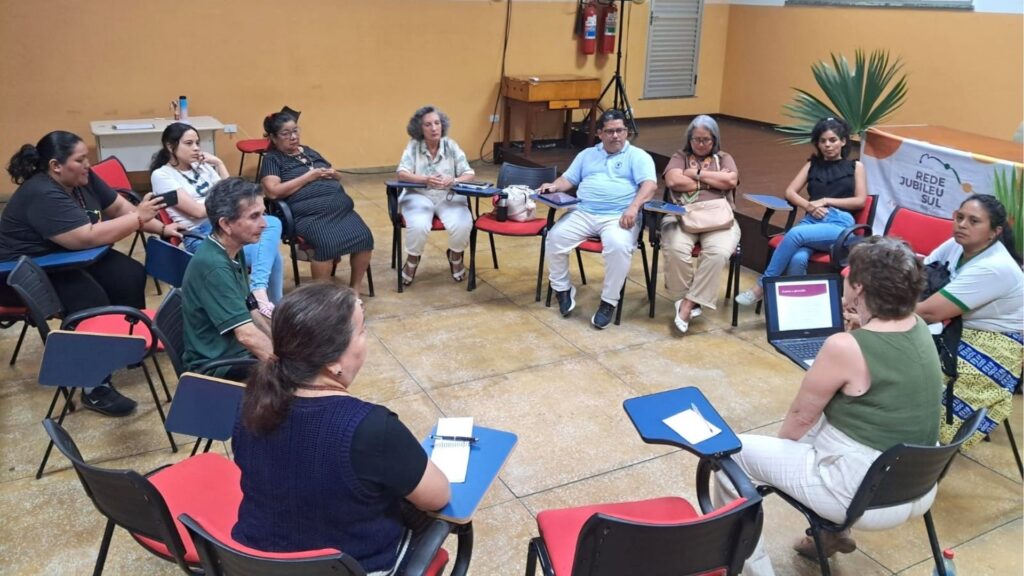 Grupo de pessoas sentadas em círculo em sala com paredes amarelas, participando de encontro da Rede Jubileu Sul.