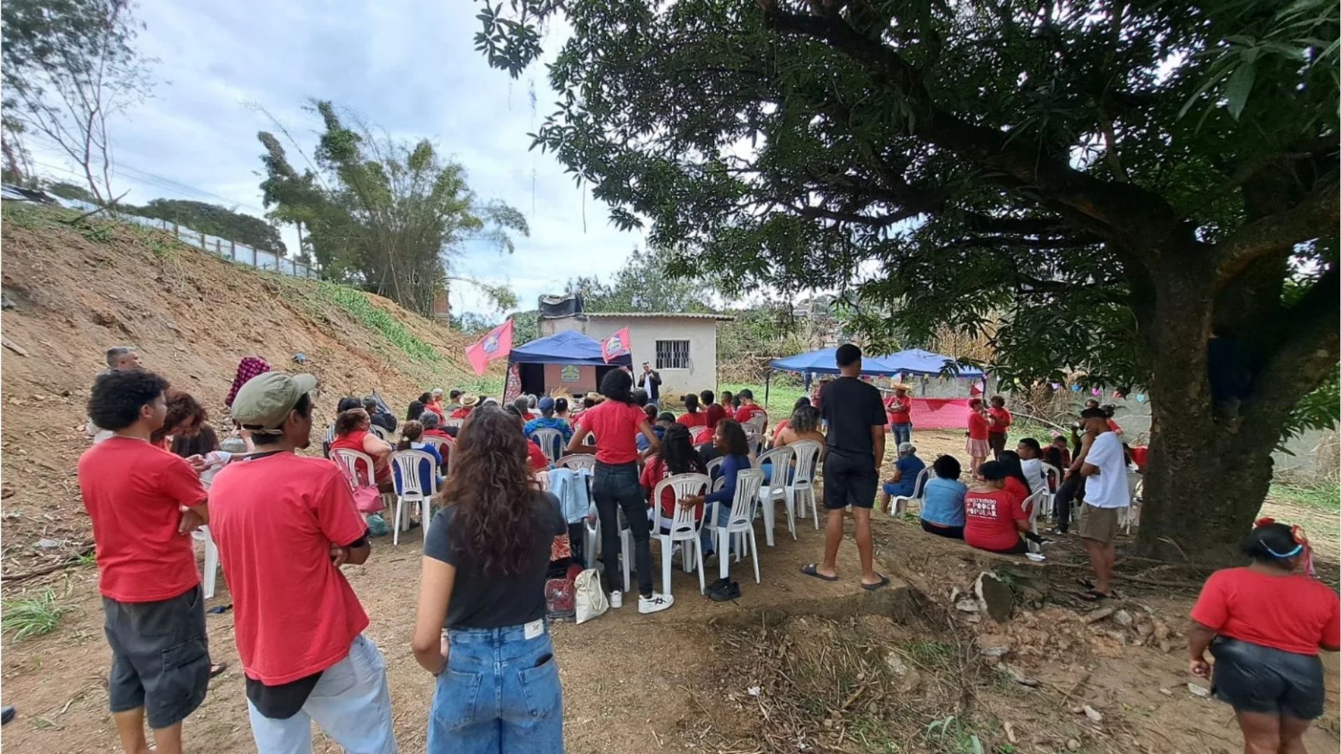 Comunidade reunida ao ar livre sob árvores, com muitas pessoas vestindo camisetas vermelhas sentadas em cadeiras brancas. Ao fundo, palhinha azul com bandeiras vermelhas e uma pequena construção branca, num ambiente rural com encosta de terra à esquerda.