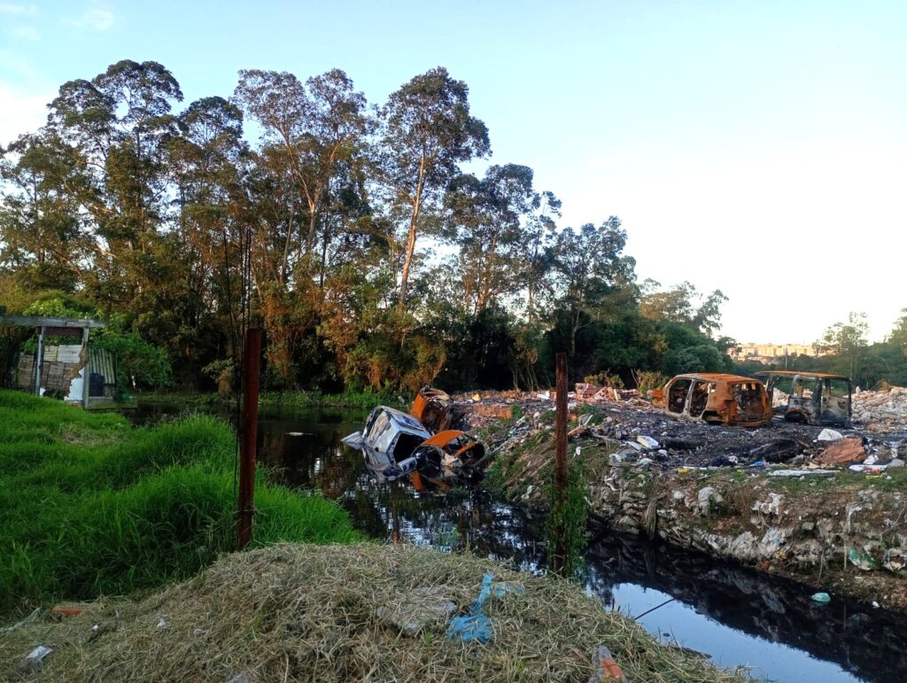Paisagem de descarte ambiental com carros queimados e entulho espalhados às margens de um rio poluído, cercado por vegetação nativa e uma pequena estrutura de madeira à esquerda.