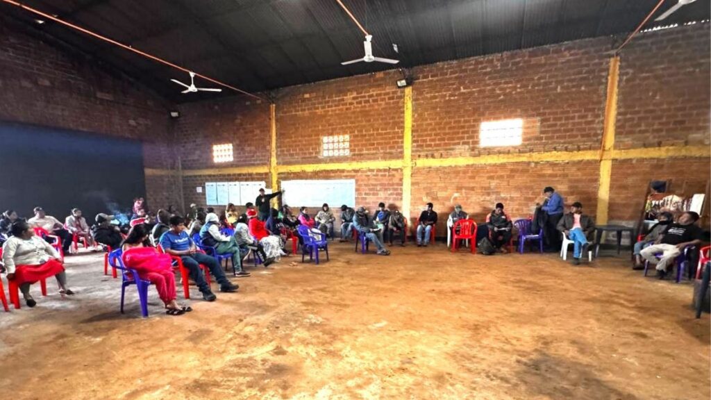 Assembleia indígena Ava Guarani reunida em grande salão com paredes de tijolos expostos e piso de terra. Participantes sentados em cadeiras plásticas coloridas formando um círculo, enquanto uma pessoa aponta para quadros brancos na parede durante apresentação ou discussão coletiva.