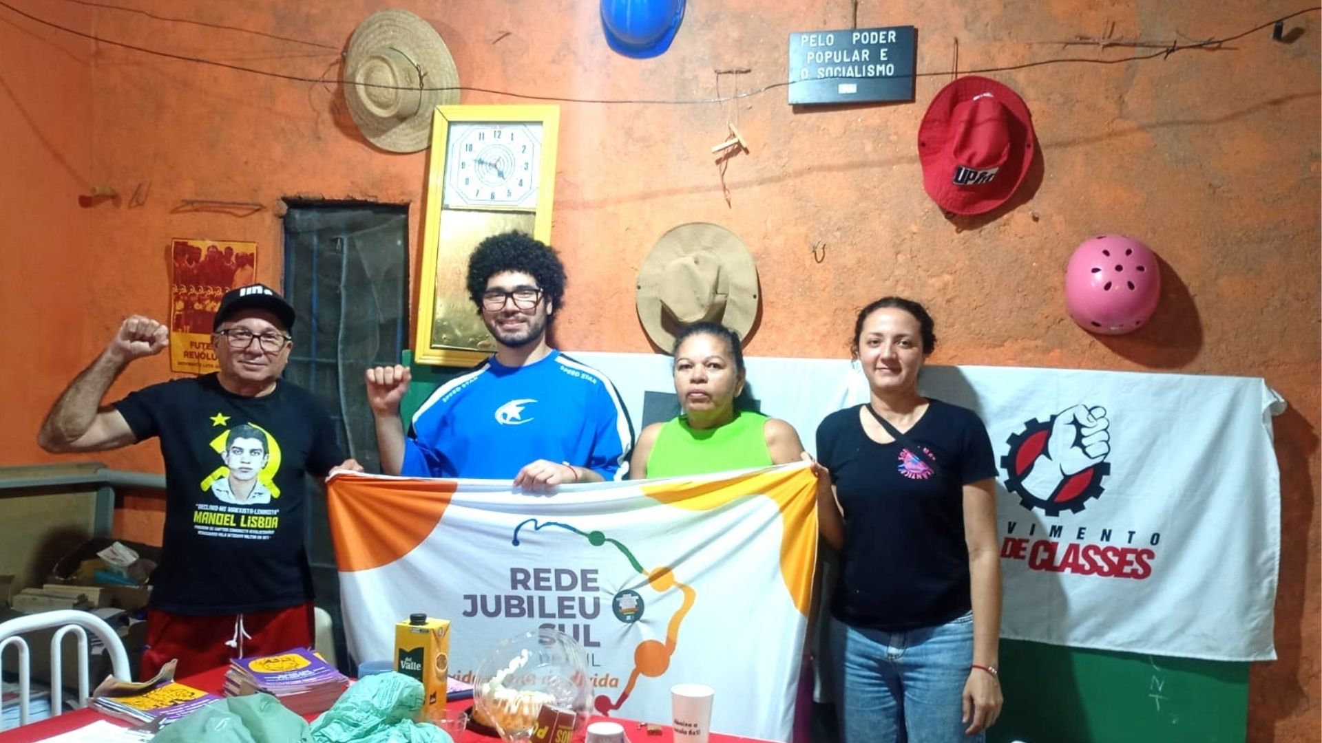 Quatro pessoas posam com bandeiras da Rede Jubileu Sul Brasil e do Movimento de Classes em um ambiente com paredes laranja decoradas com chapéus, capacetes e um letreiro que diz "PELO PODER POPULAR E O SOCIALISMO". Um homem à esquerda usa camiseta com a imagem de Manoel Lisboa e levanta o punho.
