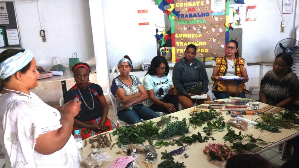 Mulheres participam de oficina com ervas e plantas medicinais dispostas sobre uma mesa, enquanto uma instrutora explica o processo. Ao fundo, cartaz colorido com mensagens sobre combate ao trabalho infantil e reconhecimento do trabalho produtivo.