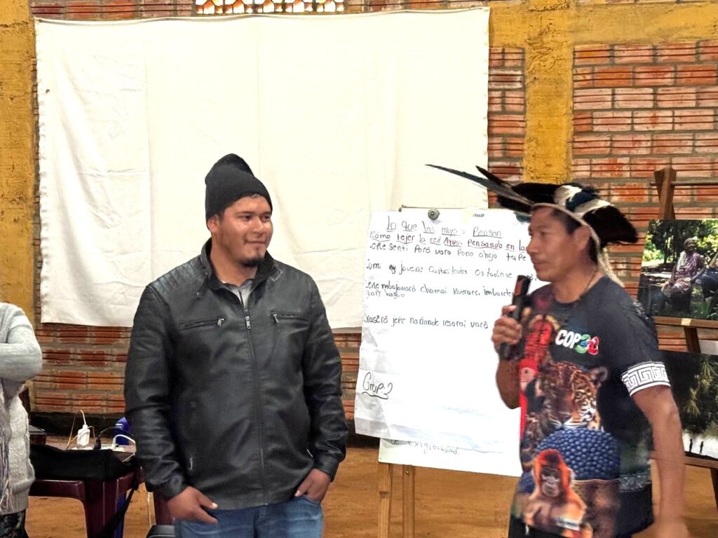 ois homens indígenas em apresentação. À direita, um porta-voz com cocar tradicional e camiseta COP30 segura microfone, enquanto à esquerda outro jovem escuta atentamente. Ao fundo, painel branco com anotações manuscritas sobre organização da rede Aruva e demandas territoriais.
