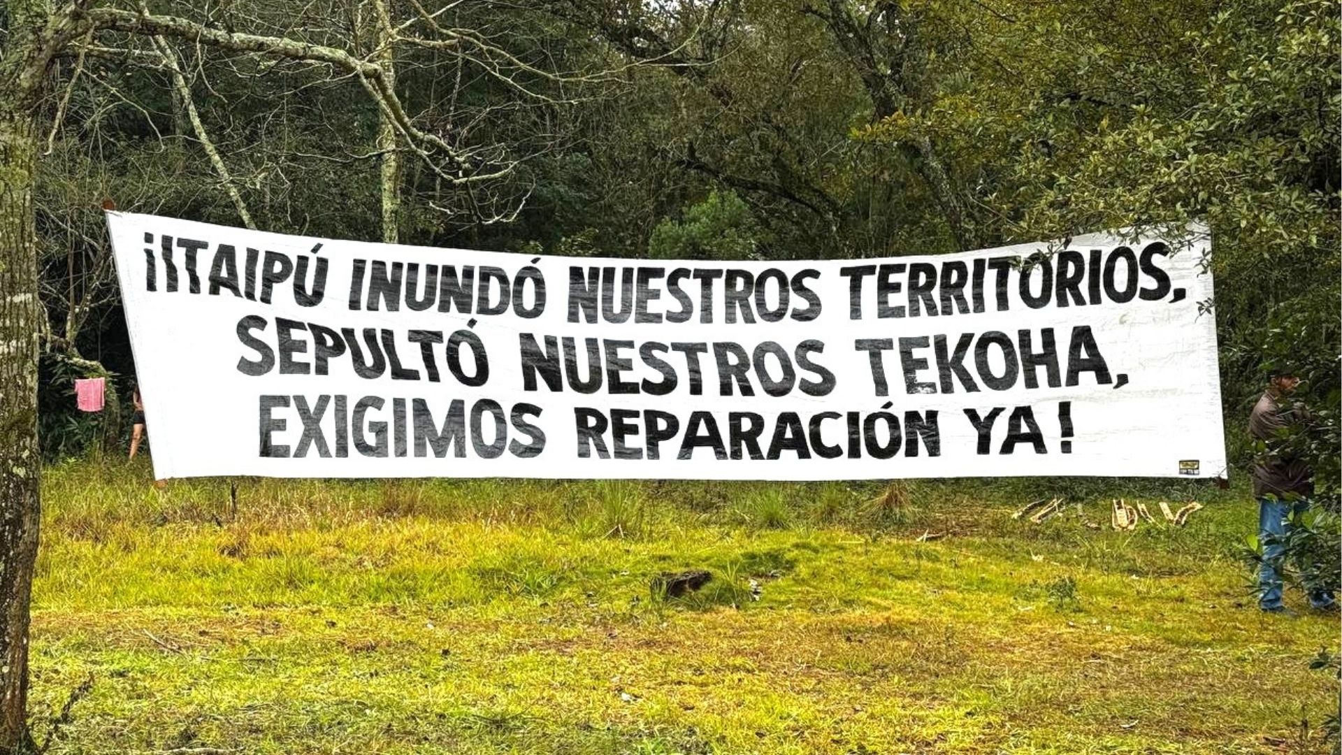 Faixa branca estendida entre árvores com mensagem em espanhol pintada à mão: "¡ITAIPÚ INUNDÓ NUESTROS TERRITORIOS, SEPULTÓ NUESTROS TEKOA, EXIGIMOS REPARACIÓN YA!" – denunciando os impactos da usina hidrelétrica sobre territórios indígenas e exigindo reparação.