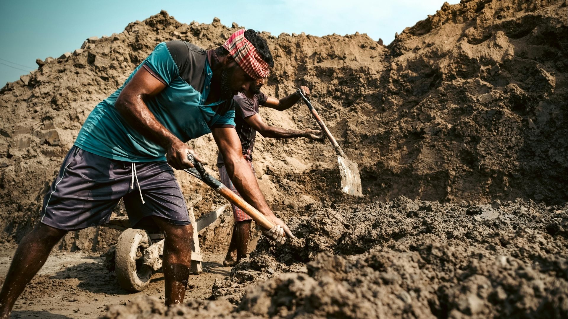 Dois trabalhadores realizando trabalho braçal pesado com pás em um terreno coberto de terra e lama, sob sol forte.