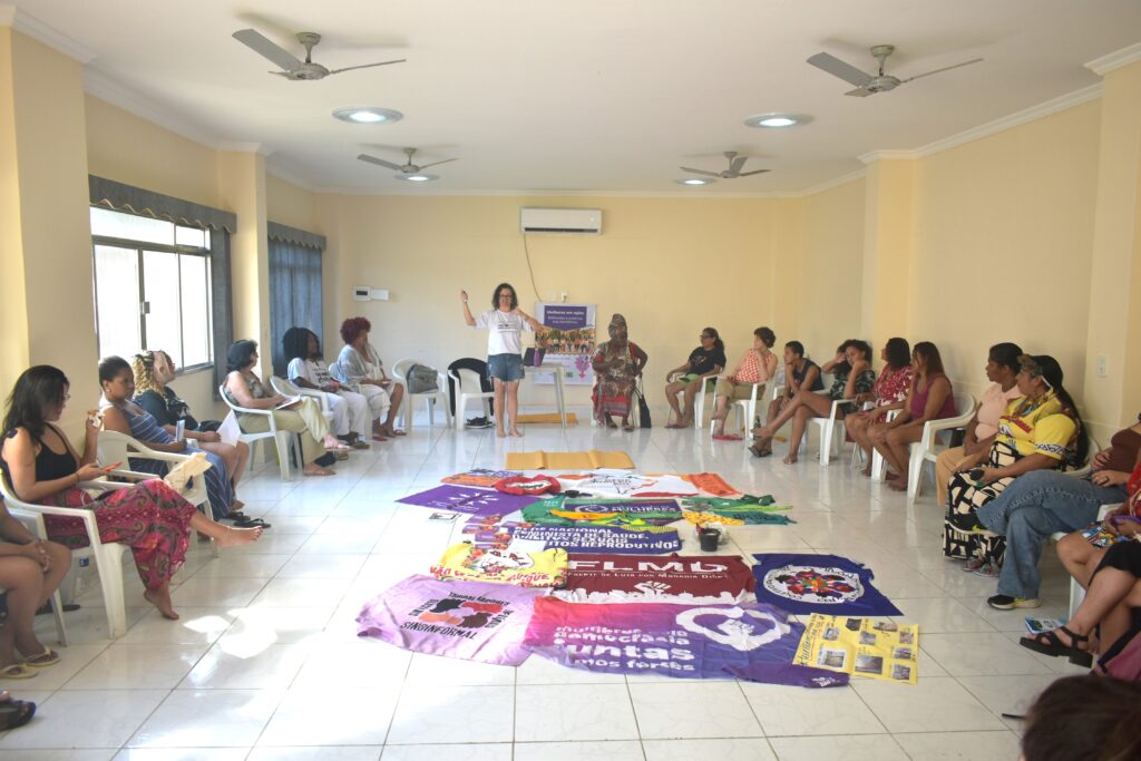 Encontro de mulheres sentadas em cadeiras plásticas formando um semicírculo numa sala clara com ventiladores no teto; uma palestrante está de pé ao centro, gesticulando, enquanto outra mulher sentada observa. No chão, entre as participantes, estão espalhadas camisetas coloridas de movimentos feministas e de direitos reprodutivos, como "Mulheres pela Democracia", "Trabalhadoras Sindinformal" e "Rede Nacional Feminista de Saúde".