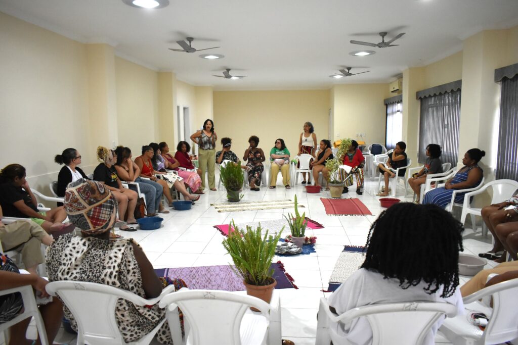 Encontro de mulheres sentadas em cadeiras plásticas formando um grande círculo numa sala ampla com ventiladores no teto. No centro do espaço, tapetes coloridos e vasos com plantas ornamentais dividem o ambiente, enquanto duas participantes estão de pé conduzindo a roda de conversa.