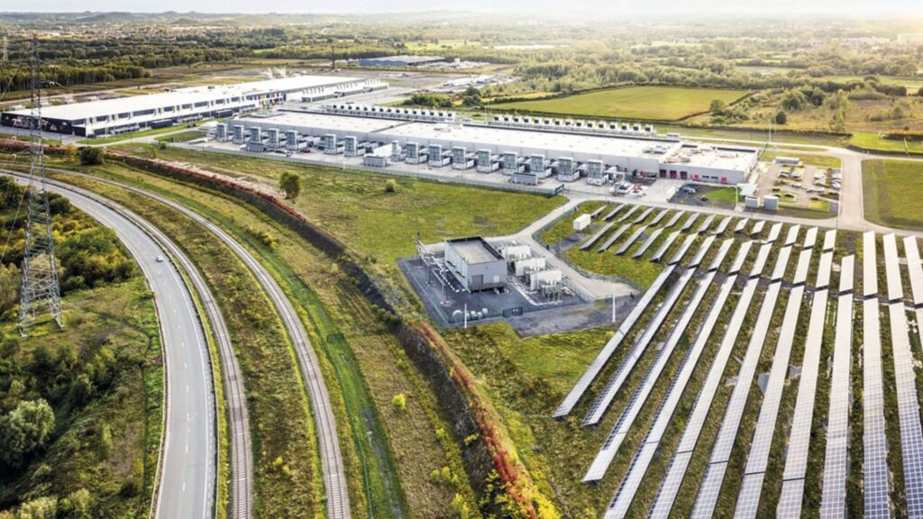 Vista aérea de um complexo industrial composto por grandes galpões brancos, possivelmente data centers, acompanhado por extensas fileiras de painéis solares fotovoltaicos instalados no solo. Ao lado da infraestrutura, observam-se trilhos ferroviários, uma estrada asfaltada e torres de transmissão elétrica cortando o campo verdejante sob luz diurna suave.