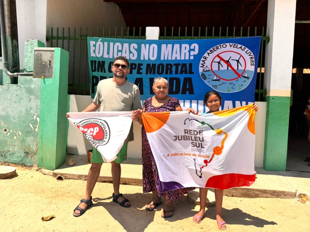 Três pessoas seguram bandeiras de organizações sociais em frente a um muro pintado de verde. Ao fundo, uma grande faixa azul exibe o slogan "EÓLICAS NO MAR? UM CAMINHO MORTAL PARA A VIDA" e o logotipo da campanha "Mar Aberto Velas Livres", que mostra turbinas eólicas com um símbolo de proibição sobre elas. As duas bandeiras à frente pertencem ao Conselho Popular (CP) e à Rede Jubileu Sul Brasil, cujo lema é "A vida acima da dívida".
