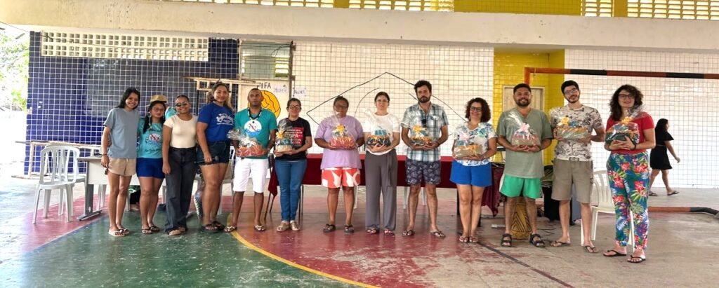 Fotografia panorâmica de um grupo de aproximadamente quinze pessoas posando em pé, lado a lado, dentro de um pavilhão coberto com piso pintado. A maioria dos participantes segura cestas básicas contendo alimentos variados, como frutas e verduras, embrulhadas em plástico transparente. O cenário ao fundo revela paredes revestidas com azulejos brancos e azuis, além de mesas e cadeiras plásticas brancas típicas de espaços comunitários ou escolares