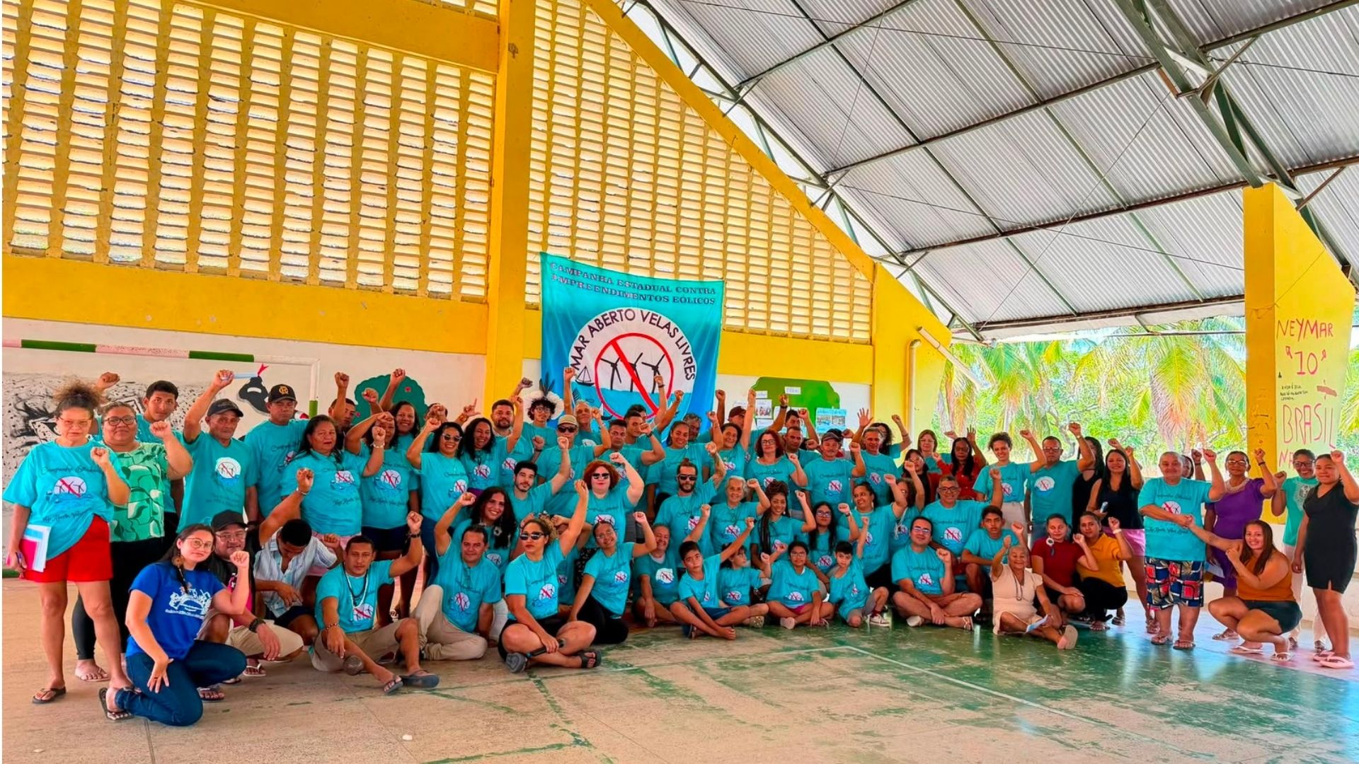 Grupo numeroso de pessoas vestindo camisetas azuis tirando foto coletiva em um pavilhão com estrutura amarela. Muitos participantes erguem o punho fechado em sinal de protesto ou solidariedade. Ao centro, destacam-se duas faixas: uma azul com a inscrição 'CAMINHA POR UM BRASIL LIVRE DE ENERGIAS FÓSSEIS' e um símbolo proibitivo sobre turbinas eólicas; outra, à direita, exibe a frase 'NEYMAR 10 BRASIL'. O ambiente sugere um evento político ou de mobilização social ao ar livre