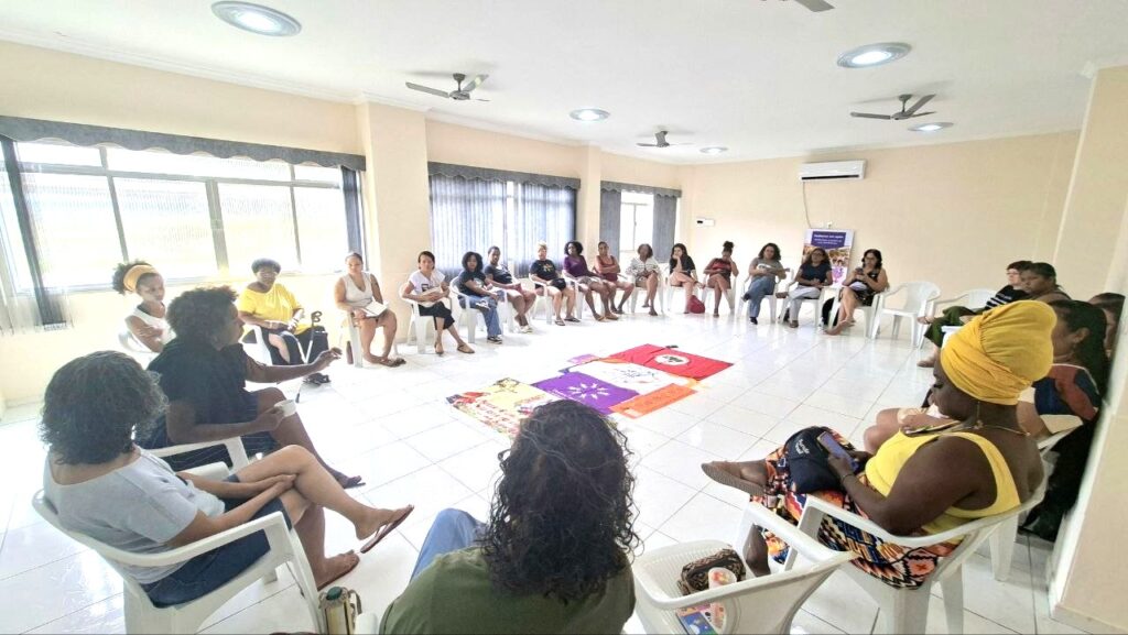 Roda de educação popular com mulheres sentadas em cadeiras plásticas formando um círculo amplo numa sala clara. No centro do chão, camisetas coloridas de movimentos sociais estão estendidas sobre o piso branco, enquanto as participantes se escutam atentas durante o encontro.